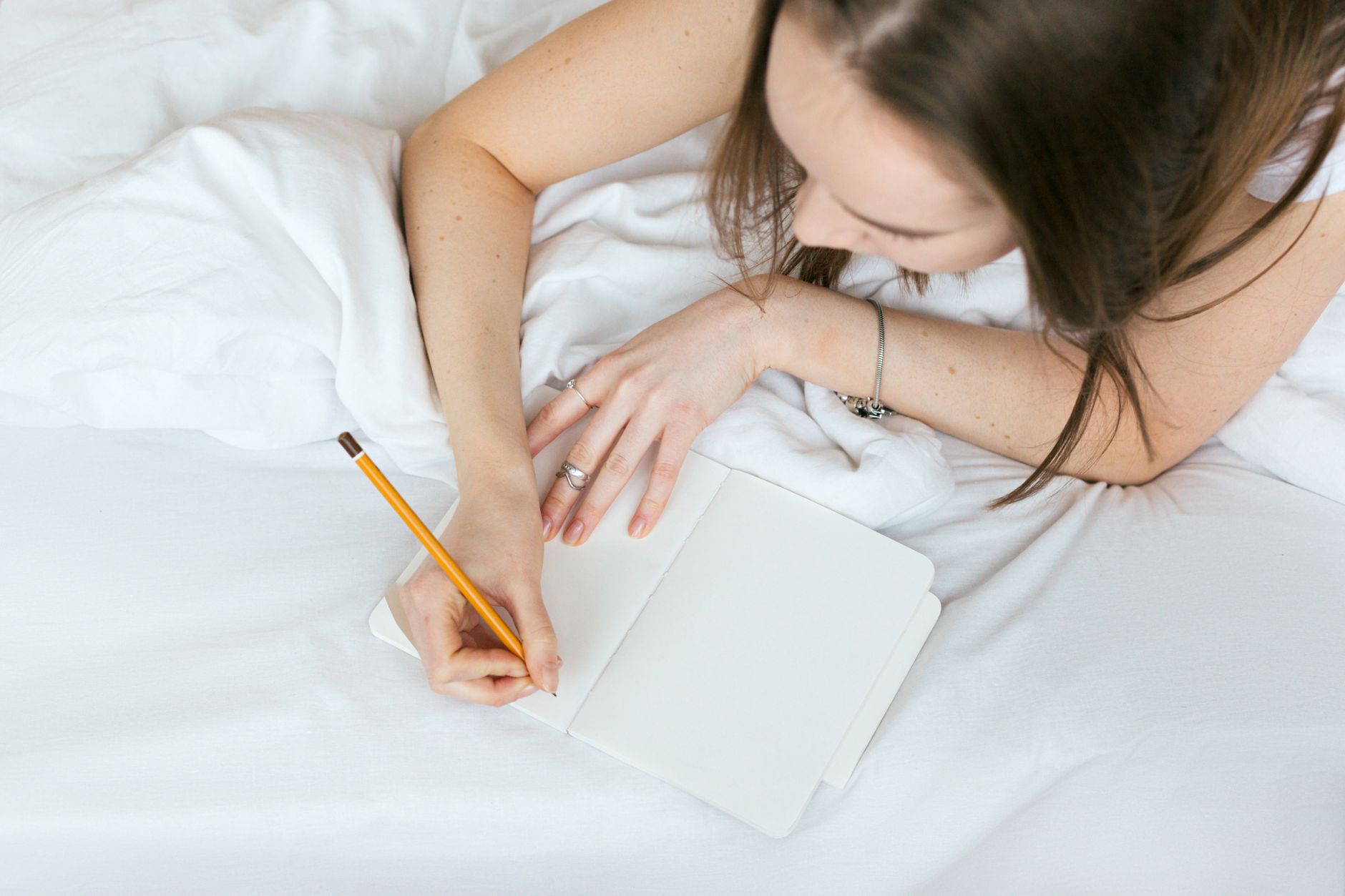 Woman relaxing on a bed while writing in a journal with a pencil.